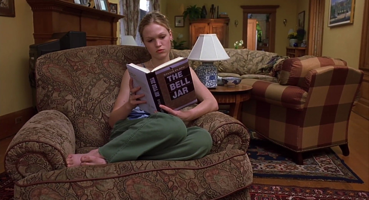 Film Still of Katniss Everdeen from 10 Things I hate about you reading a copy of Sylvia Plath's The Bell Jar whilst sat in an arm chair in her family home.