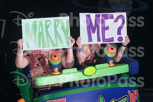 Photopass photo of Chelsea and Lacey holding the "Marry" and "Me" signs on Buzz Lightyear.