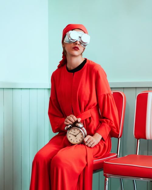 Futuristic photo of a women sitting in a bright red robe/dress with aluminum retro futuristic goggles holding a clock in front of an aqua blue background.