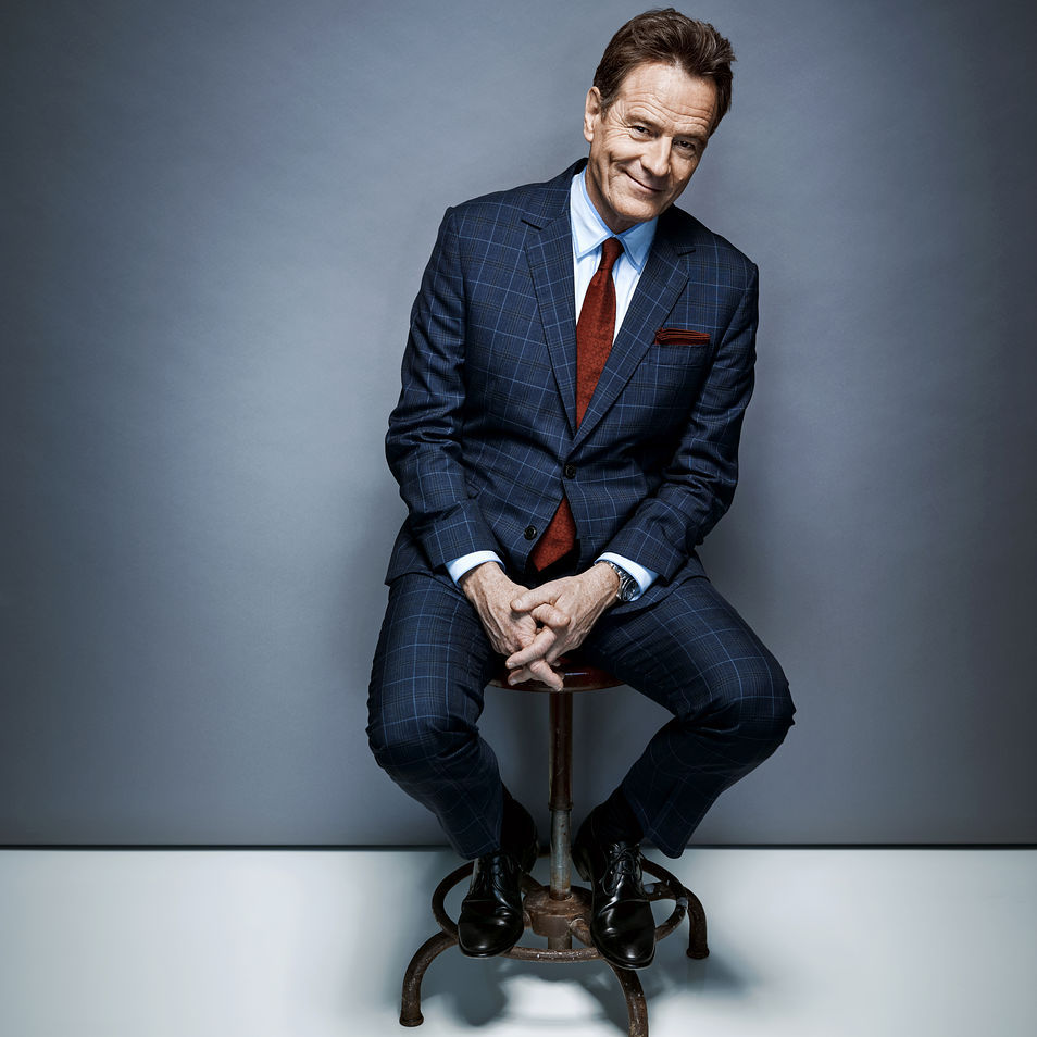 Bryan Cranston sits on a stool in front of a gray backdrop. He wears a Brooklyn Tailors navy plaid suit.