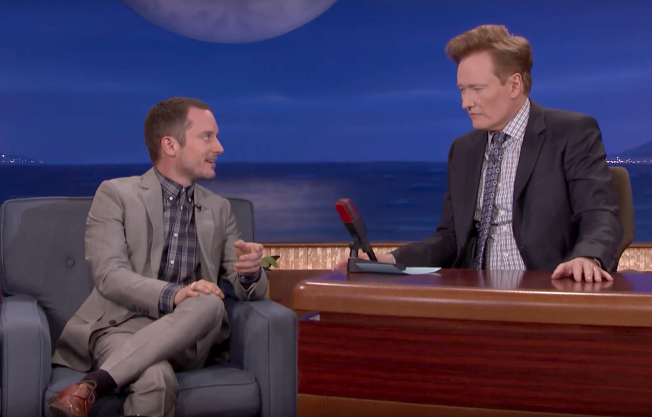 Still from Conan of Elijah Wood wearing a gray suit and blue/gray plaid shirt from Brooklyn Tailors seated next to Conan O'Brien on his late night show.